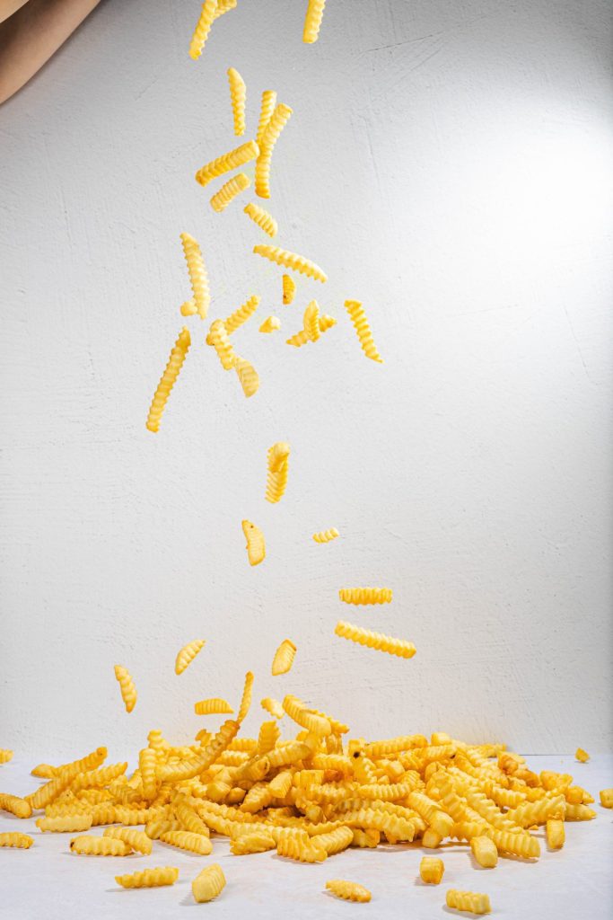 Crinkle-cut French fries falling from above and piling onto a white surface against a textured white background. The fries appear golden and crispy, captured mid-air as they cascade down.