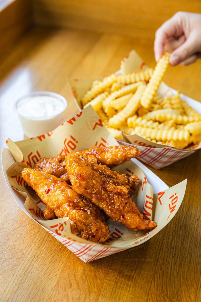 A basket of crispy, saucy chicken tenders paired with a side of golden crinkle-cut fries and a cup of dipping sauce, served on a wooden table. A hand reaches in to grab a fry.